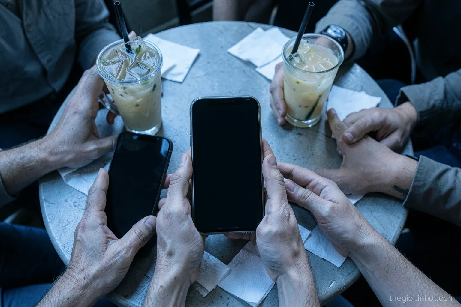 Top-down view of several hands around a cafe table each holding a phone with a blank dark screen, iced drinks and scattered napkins, cool morning light, desaturated blue palette
